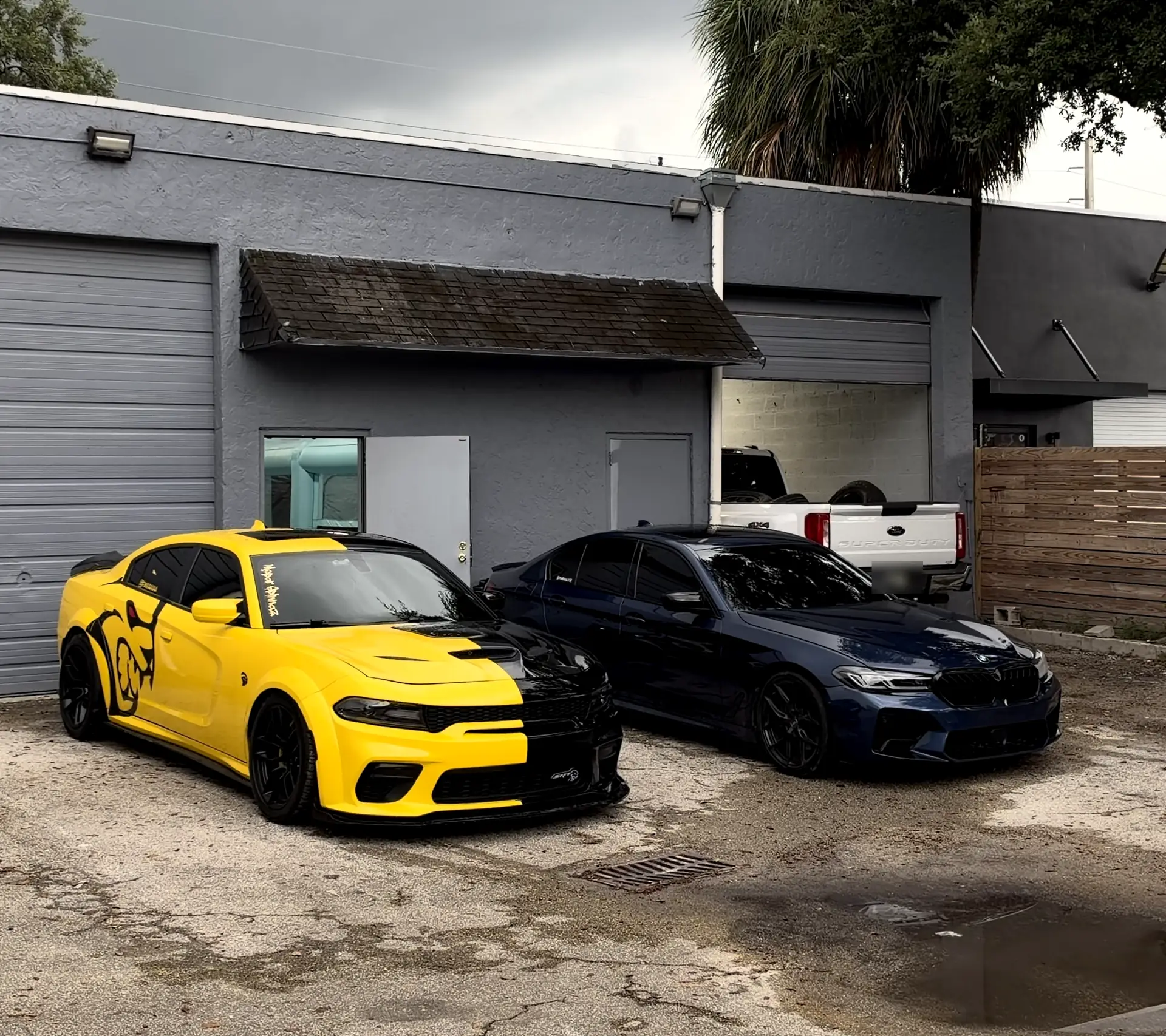 Fima’s Autoworks on SW 38th St: the grey shop building with roll-up bay doors, Fima’s yellow-and-black Dodge Charger Hellcat Widebody and a dark blue BMW M5 parked in front